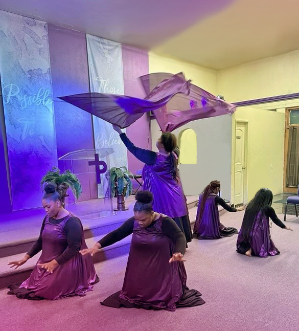 Members of the Peculiar People dance group performing in harmonious movement at Power of Praise church, with a dancer gracefully raising a purple flag while others kneel and extend their arms in worship.